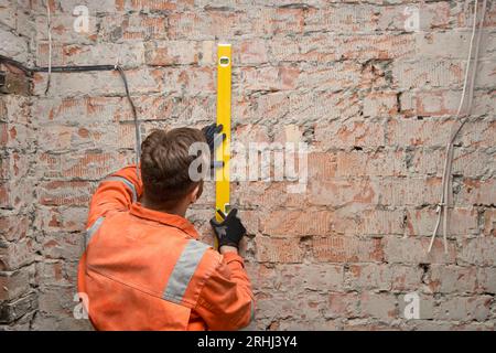 Bauarbeiter, der mit einer Wasserwaage eine senkrechte Linie auf eine Ziegelwand in Innenräumen zeichnet. Mann trägt orangefarbene Overalls. Stockfoto