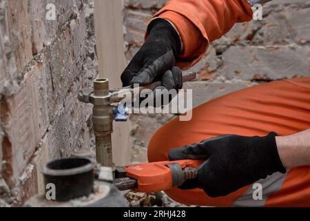 Installateur repariert undichten Wasseranschluss vor Ort, trägt Handschuhe und orangefarbene Overalls. Stockfoto