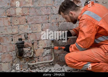 Klempner repariert undichten Wasseranschluss. Klempner trägt orangefarbene Overalls und Handschuhe. Stockfoto