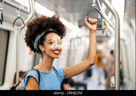Positive moderne junge marokkanische Frau mit Afro-Haarband lächelnd und wegschauend, während sie steht und den Griff während der Reise in Unschärfe hält Stockfoto