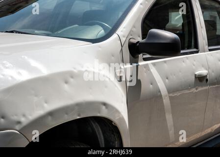 Beschädigtes weißes Auto mit vielen Dellen, die an einem sonnigen Tag nach einem Hagelsturm auf der Stadtstraße geparkt wurden Stockfoto