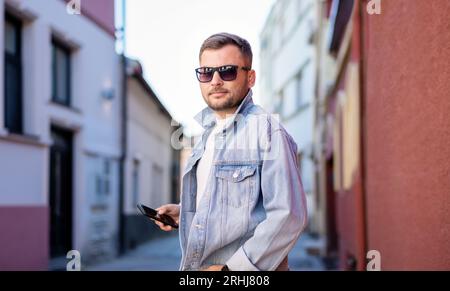 Trendiger junger Erwachsener in Sonnenbrille und Jeansjacke, der auf der Straße steht und das Handy in der Hand hält. Stockfoto