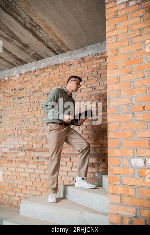 Ganze Länge des Maklers mit Faltschachtel, der auf Treppen im Haus steht, mit unvollendetem Innenraum Stockfoto