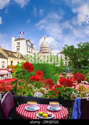 Paris Café Restaurant Coffee Cups View Vista Montmartre Frühlingssommerblumen Kaffee für zwei und kleine Kuchen Makronen auf französisch karierter Tischdecke mit Blumenblick. Französische dreifarbige Flagge mit Blick auf den Place du Tertre, Sacre Coeur hinter Montmartre Paris Frankreich Stockfoto