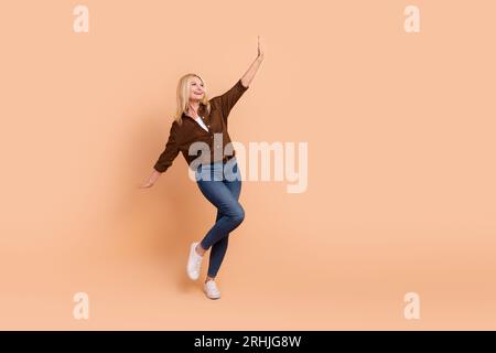 Ganzkörperfoto einer überfreudigen Mittelalterfrau, die Hände hochhebt und begeistert einen isolierten beigefarbenen Hintergrund tanzt Stockfoto