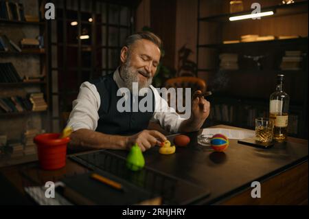 Lustig eleganter älterer Geschäftsmann raucht Zigarre und spielt Spielzeug am Arbeitstisch Stockfoto