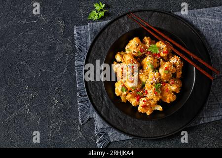 Knusprige orangefarbene Blumenkohlröschen, bestreut mit Sesamsamen, Chiliflocken und Petersilie in schwarzer Schüssel auf Betontisch mit Stäbchen, horizontal VI Stockfoto