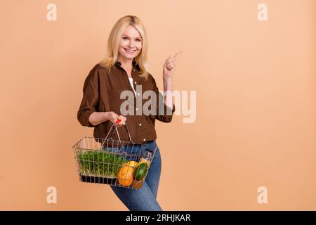 Foto einer jungen Frau blonde Geschäft Hypermarkt tragen braune Hemd halten Korb Bio-Lebensmittel direkt Finger Neuheit isoliert auf beigem Hintergrund Stockfoto