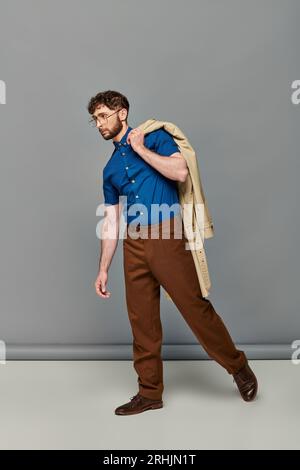 Bärtiger Mann in Brille mit Trenchcoat, grauer Hintergrund, stilvolles Modell, durchgehend, elegant und lässig Stockfoto