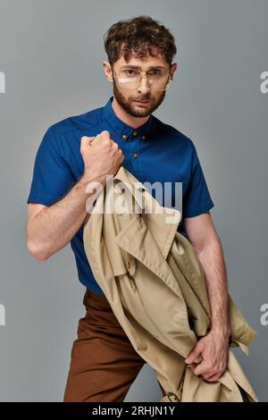 Stilvoller Mann in Brille mit Trenchcoat auf grauem Hintergrund, Blick auf die Kamera, elegant und lässig Stockfoto