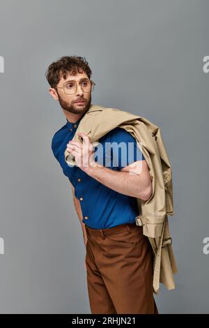 Bärtiger Mann in Brille mit Trenchcoat, grauer Hintergrund, männliches Model schaut weg, elegant lässig Stockfoto