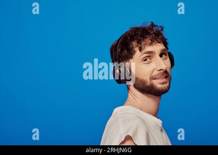 Positiver Mann mit schnurlosen Kopfhörern, der Musik auf blauem Hintergrund, Männermode und Audio hört Stockfoto