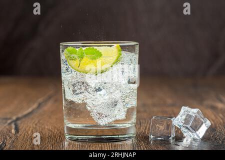 Kalt, erfrischend in der Hitze, prickelndes Wasser im Glas mit Eiswürfeln, Limettenkeil und Minzblättern. Dunkler hölzerner Bartheke Hintergrund. Künstliche Klimaanlage Stockfoto