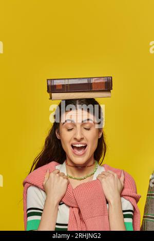 Aufgeregte Frau mit geschlossenen Augen stehend mit Buch auf Kopf, gelber Hintergrund, Studentenleben, jung Stockfoto