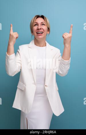 Fröhliche, hübsche, blonde, junge europäische Frau in einer weißen Jacke und Kleid zeigt mit dem Finger auf die Werbung Stockfoto