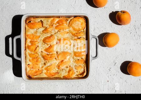Clafoutis mit Aprikose - Sommertorte in einer Auflaufform und frische Aprikosen auf dem hellen Tisch, Blick von oben Stockfoto