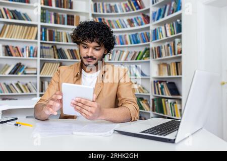 Junger erfolgreicher Student, der in der akademischen Universitätsbibliothek sitzt, lächelnder Mann, der Tablet-Computer hält, hispanischer Mann, der die Studien-App verwendet. Stockfoto