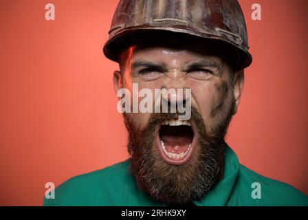 Close-up-Porträt schreiender Bauarbeiter im Schutzhelm. Wütender Erbauer im Schutzhelm. Schreiender bärtiger Mann in Bauuniform und Schutzhelm Stockfoto