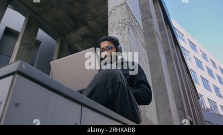 Indian Guy Universitätsstudent in Kopfhörer Hören Sie sich ein Business-Webinar oder einen Fernkurs an, der im Freien studiert. Arabischer Mann Geschäftsmann männlicher Freiberufler in der Stadt Stockfoto