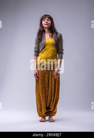 Portrait einer erwachsenen Frau vor neutralem Hintergrund, in warmen Gelb- und Beigetönen gekleidet, mit Puffhose und Barfuß. Ihr Gesicht drängt Stockfoto