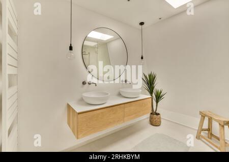 Ein Badezimmer mit zwei Waschbecken und einem Spiegel an der Wand daneben ist eine Pflanze in einem Holzhocker Stockfoto