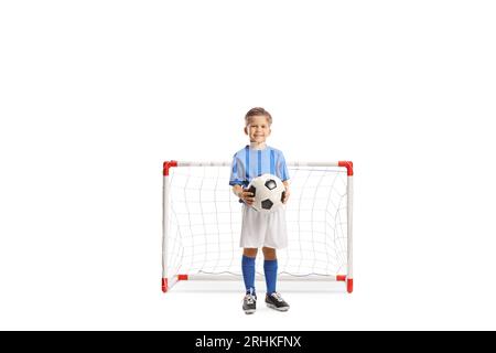 Niedlicher kleiner Junge, der einen Fußballball hält und vor einem Minitor auf weißem Hintergrund lächelt Stockfoto