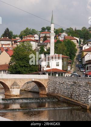 Brücke über den Fluss Miljacka mit Restaurant Inat Kuca und Minarett hinter und Hügellandschaften. Sarajevo, Bosnien und Herzegowina, 17. August 2023. Stockfoto