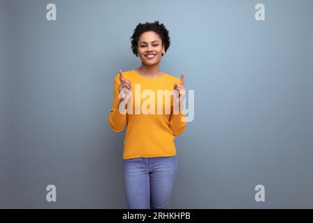 Junge lateinamerikanische Frau mit schwarzem lockigem Haar in einem gelben Pullover zeigt mit dem Zeigefinger nach oben Stockfoto