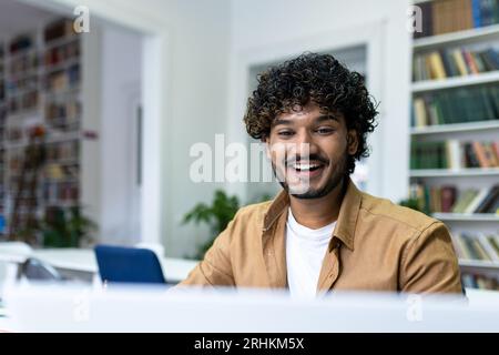 Indischer Student mit lockigen Haaren, der zwischen Büchern in Regalen sitzt, ein Mann, der sich den Videokurs ansieht, der in Notizbüchern schreibt, lächelt zufrieden. Stockfoto