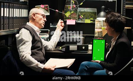 Detectives bewerten Greenscreen-Display, um zu versuchen, kriminelle Fälle zu lösen, mit Chromakey-Tablet im Polizeiarchiv. Team von Agenten, die Strategie mit isoliertem Bildschirm diskutieren, den Täter fangen. Stockfoto