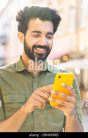 Glücklicher lächelnder bärtiger indischer Mann, der mit dem Smartphone Textnachrichten eingibt, im Internet surfen, soziale Medien, Web-App surfen, online im Freien chatten. Guy Tourist zu Fuß in der Stadt Straße. Vertikale Aufnahme Stockfoto