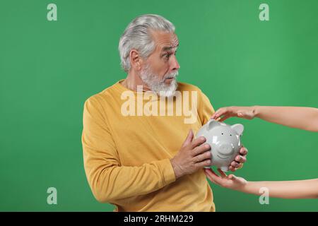 Frau, die Schweinebank von einem verwirrten Seniormann auf grünem Hintergrund nimmt. Vorsicht - Betrug Stockfoto