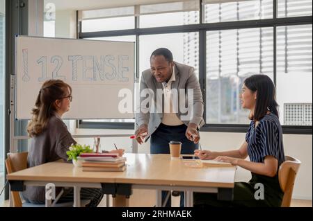 Ein professioneller afroamerikanischer englischer Lehrer diskutiert, erklärt Arbeitsaufgaben und unterrichtet asiatische Studenten im Klassenzimmer in einer Sprache Stockfoto