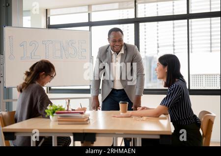 Ein professioneller und fröhlicher afroamerikanischer männlicher Englischlehrer oder -Tutor lacht und genießt das Gespräch mit asiatischen Schülern während des Unterrichts. Stockfoto