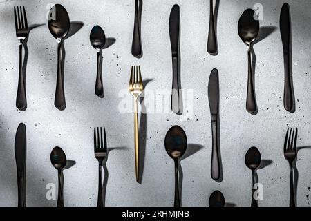 Eine goldene Gabel unter schwarzem Besteck. Neutraler grauer Betonhintergrund Stockfoto