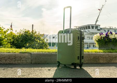 Ein großer, moderner grüner Koffer auf Rädern steht auf dem Weg vor dem Hintergrund eines weißen Passagierschiffs. Das Konzept des Sommerurlaubs und des Stockfoto