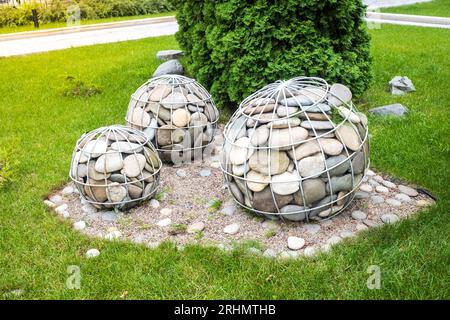 Landschaftsgestaltung. Gartenskulptur. Eisennetze in Form von Kugeln, die mit großen Steinen gefüllt sind, in einem kleinen Park mit Bänken Stockfoto