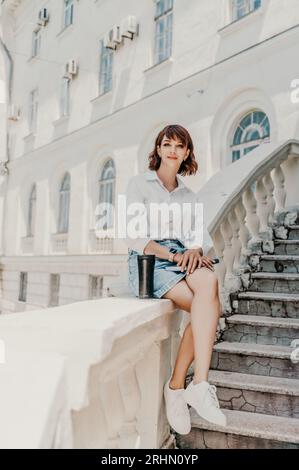 Eine Frau in einem weißen Hemd und einem blauen Jeansrock sitzt auf der Attika und hält ein blaues Notizbuch in den Händen vor dem Hintergrund des Gebäudes Stockfoto