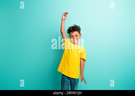 Foto eines fröhlichen Schuljungen mit braunen Haaren gekleidetes gelbes T-Shirt, das den Arm hochhebt, die Höhe auf türkisfarbenem Hintergrund isoliert Stockfoto