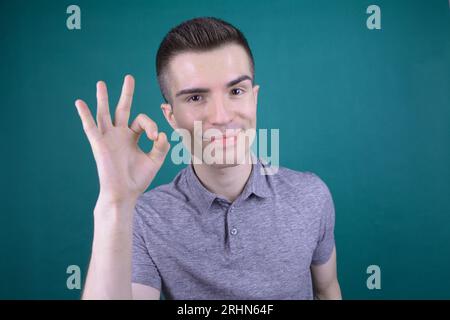 Lächelnder junger gutaussehender kaukasischer Mann in grauem T-Shirt zeigt OK-Zeichen auf isoliertem Hintergrund Stockfoto