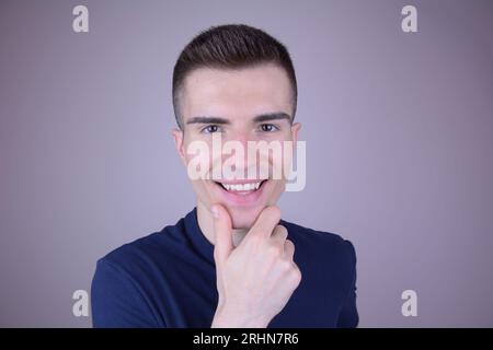 Lächelnd gutaussehender junger Mann hält Chin und schaut auf Kamera auf isoliertem Hintergrund Stockfoto