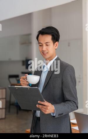 Junger asiatischer Geschäftsmann, der im Büro arbeitet und in der Hand eine Ablage hält und Morgenkaffee trinkt Stockfoto