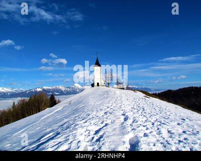 Kirche St. Primoz und Felician bei Jamnik in Gorenjska, Slowenien im Winter mit schneebedecktem Berggipfel von Storzic Stockfoto