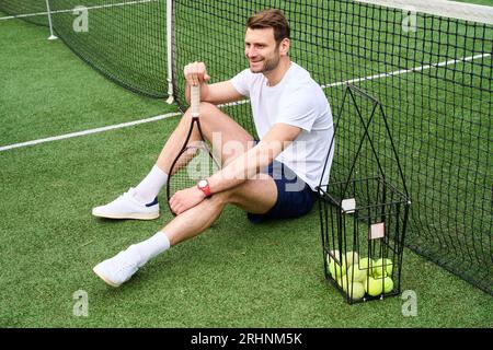 Lächelnder gutaussehender Mann, der auf dem Tennisplatz auf dem Rasen sitzt, hat einen Schläger und einen Korb mit Bälle Stockfoto