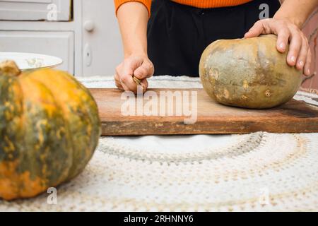 Vorderansicht einer unkenntlich gebliebenen kaukasierin mit Messer und Kürbis auf dem Schneidebrett auf dem Tisch mit rohen Kürbissen, Zubereitung von Veganer und Vegetar Stockfoto