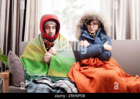 Trauriges Paar mittleren Alters in warmen Kleidern, das auf dem Sofa in ihrem Wohnzimmer kalt wird Stockfoto