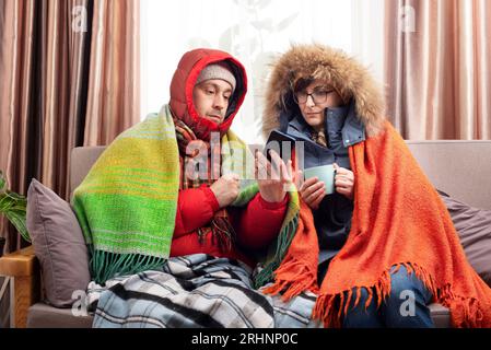 Mittelalte Familie in Parkas, die Kaffee auf dem Sofa trinkt, während sie das Smartphone in ihrem kalten Wohnzimmer benutzt Stockfoto