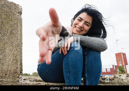 Vorderansicht der jungen lateinischen Frau draußen an einem kalten und windigen Morgen, glücklich sitzend und lächelnd mit ausgestrecktem Arm, der auf die Kamera schaut, Life Studies Co Stockfoto