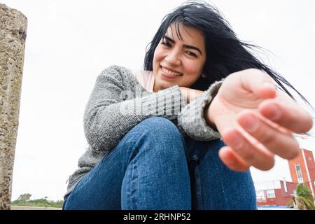 Vorderansicht der jungen lateinischen Frau draußen an einem kalten und windigen Morgen, glücklich sitzend und lächelnd mit ausgestrecktem Arm, der auf die Kamera schaut, Life Studies Co Stockfoto