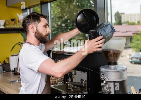 Seitenansicht des Barista in der Schürze, der Kaffeebohnen in die Mühle gießt, während er im Coffee Shop arbeitet Stockfoto